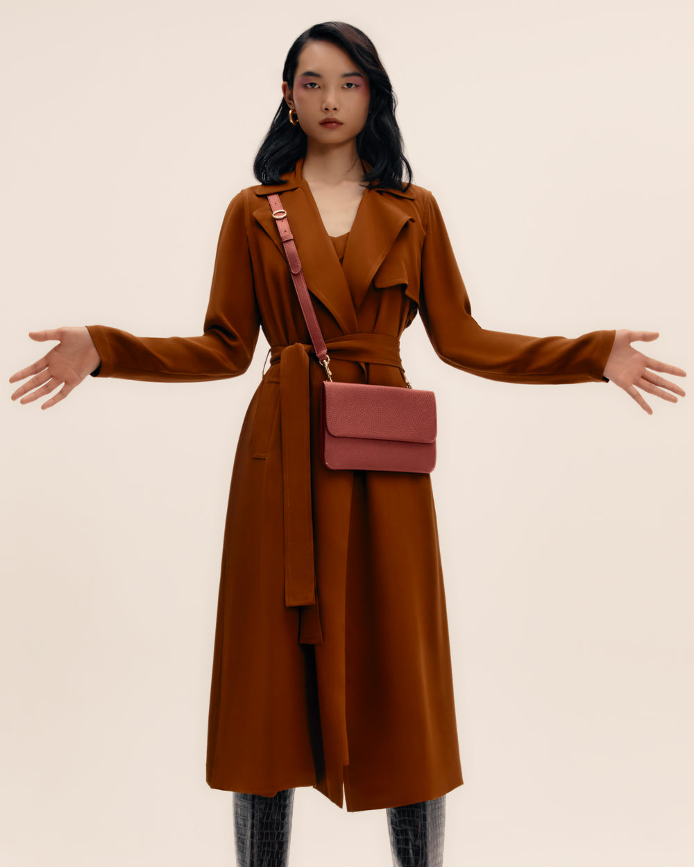 Person wearing long coat, holding arms out, with small bag across shoulder.