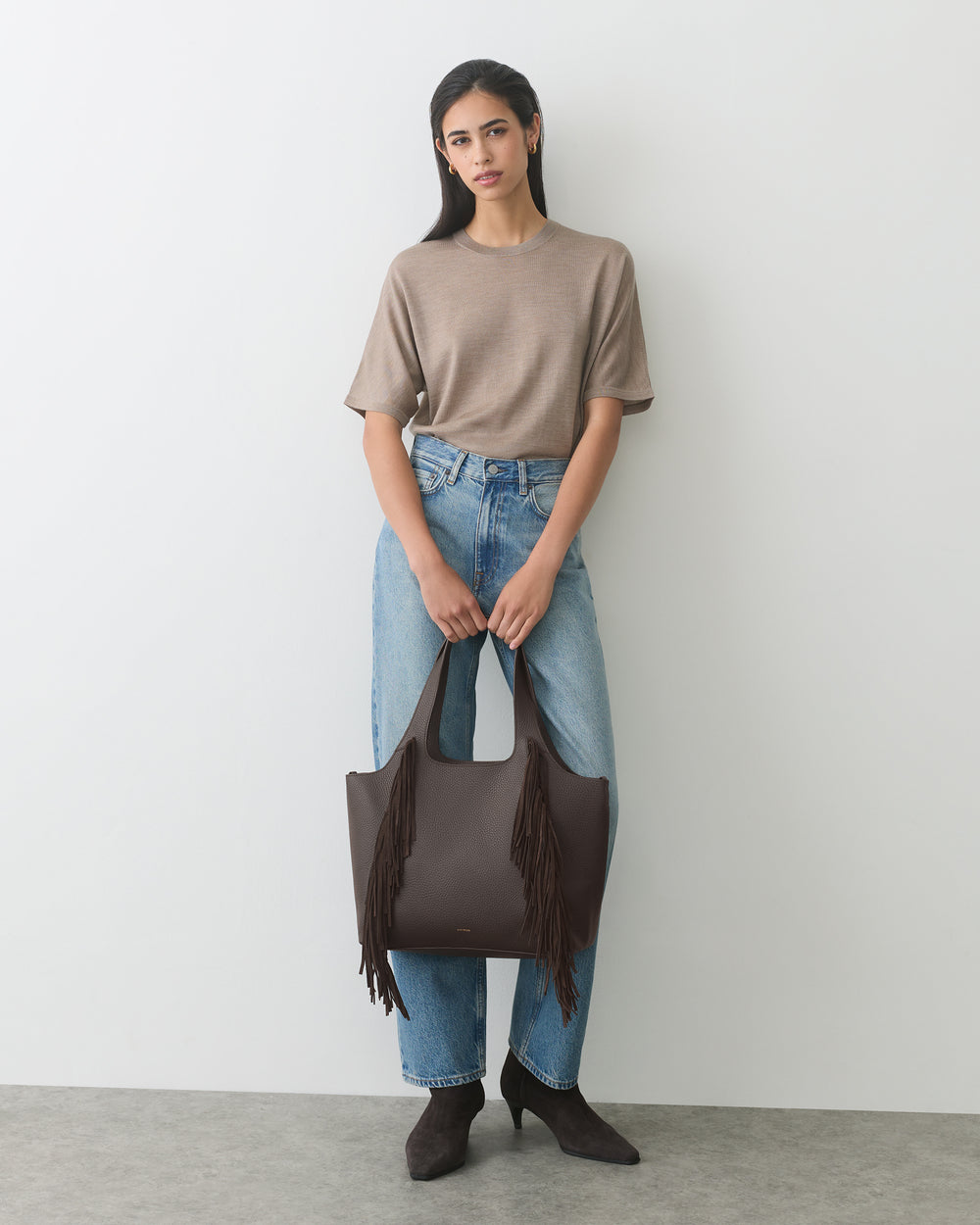 Person holding a fringed tote bag wearing a t-shirt, jeans, and ankle boots.