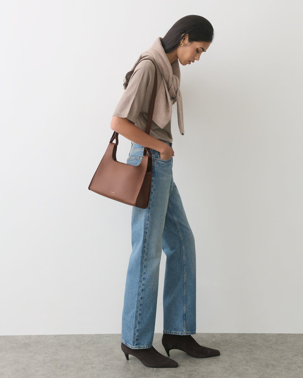 Person standing with sweater draped over shoulders, holding a handbag, wearing jeans and boots.