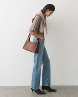 Person standing with sweater draped over shoulders, holding a handbag, wearing jeans and boots.