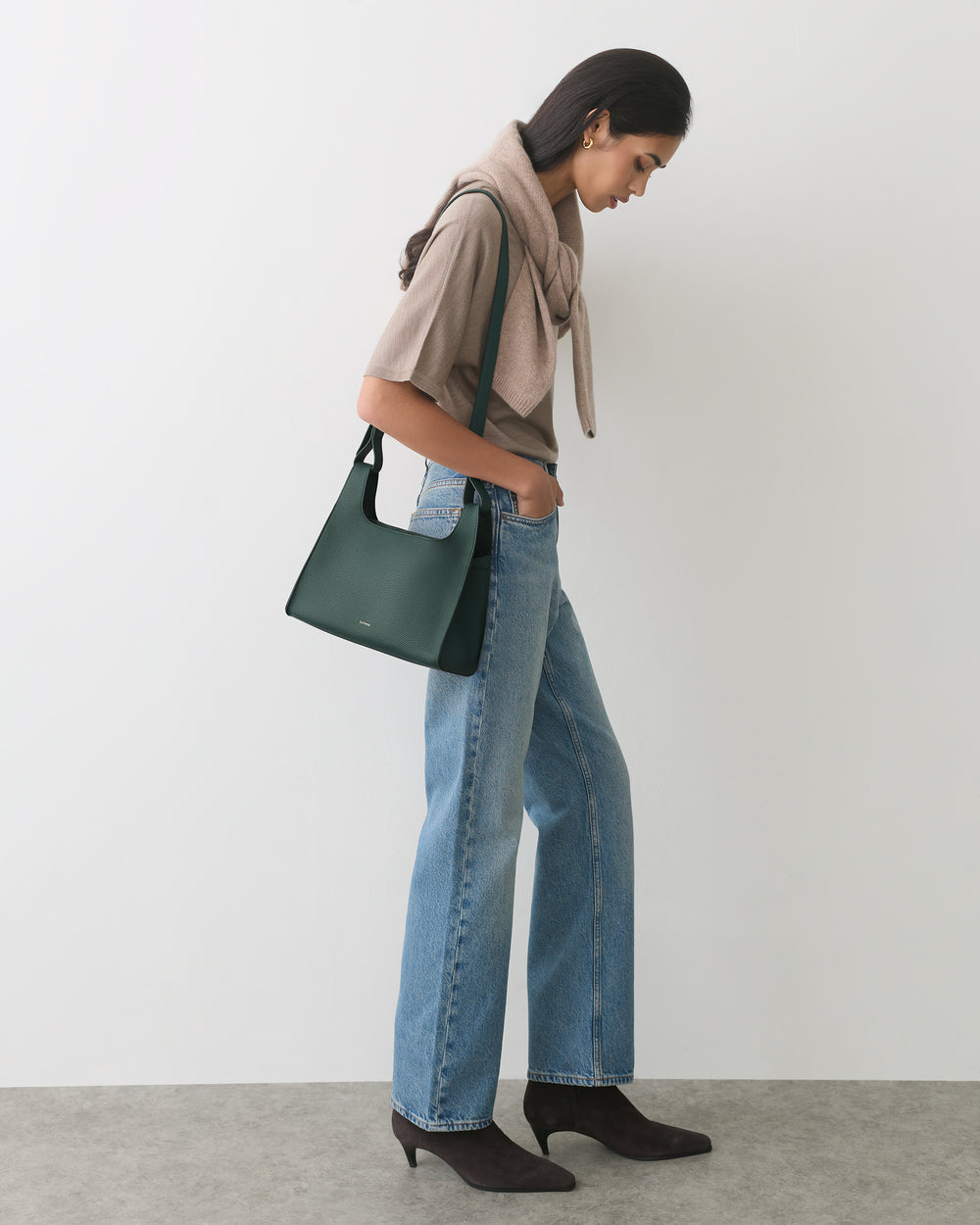 Person in a casual outfit with shoulder bag, scarf around neck, and boots, standing sideways.