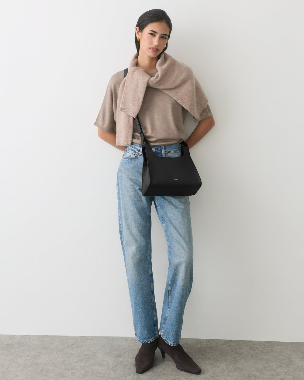 Person standing, wearing jeans and a cape, holding a black handbag.