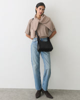 Person standing, wearing jeans and a cape, holding a black handbag.