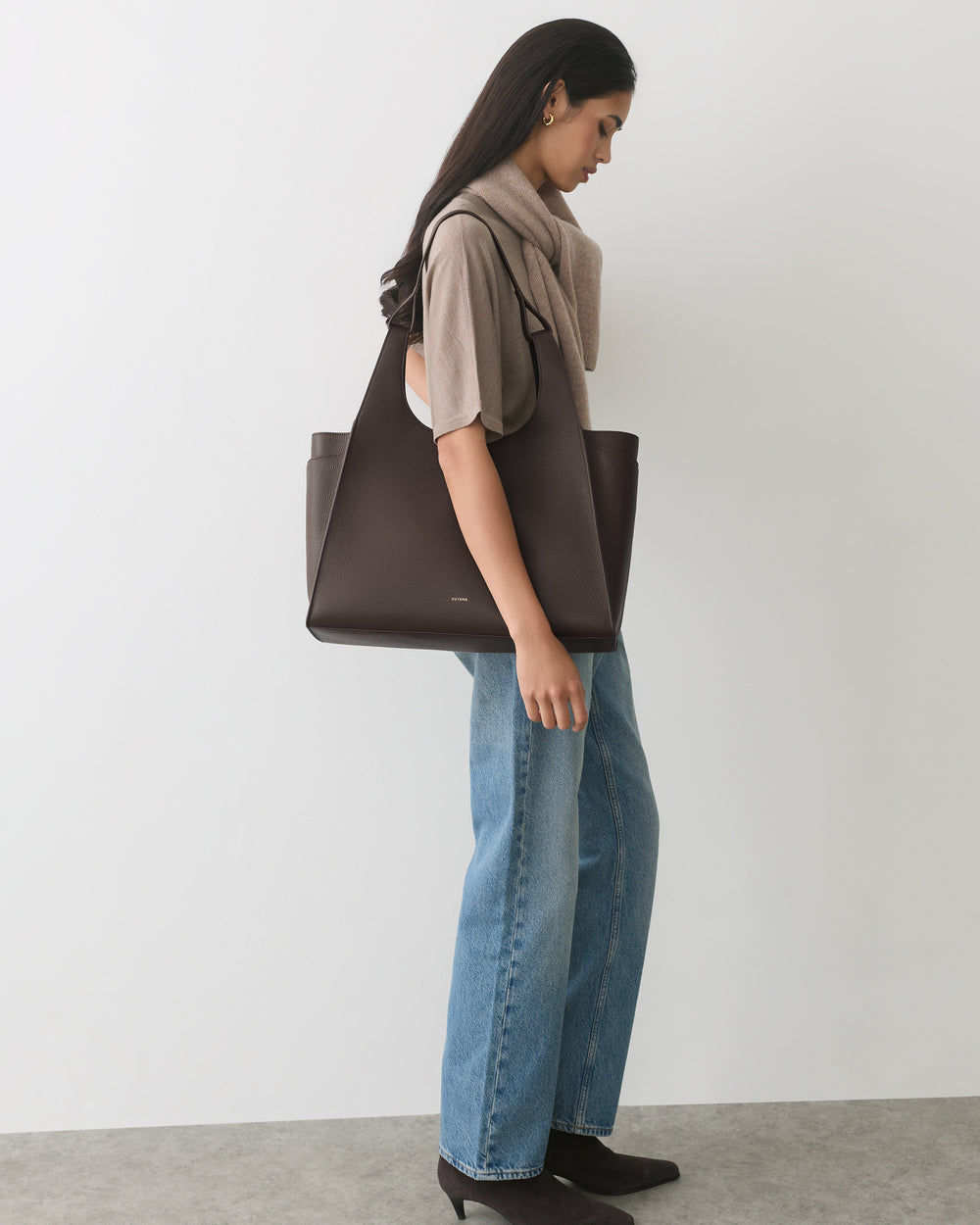 Person standing with large tote, wearing jeans and boots, with a scarf draped over shoulders.