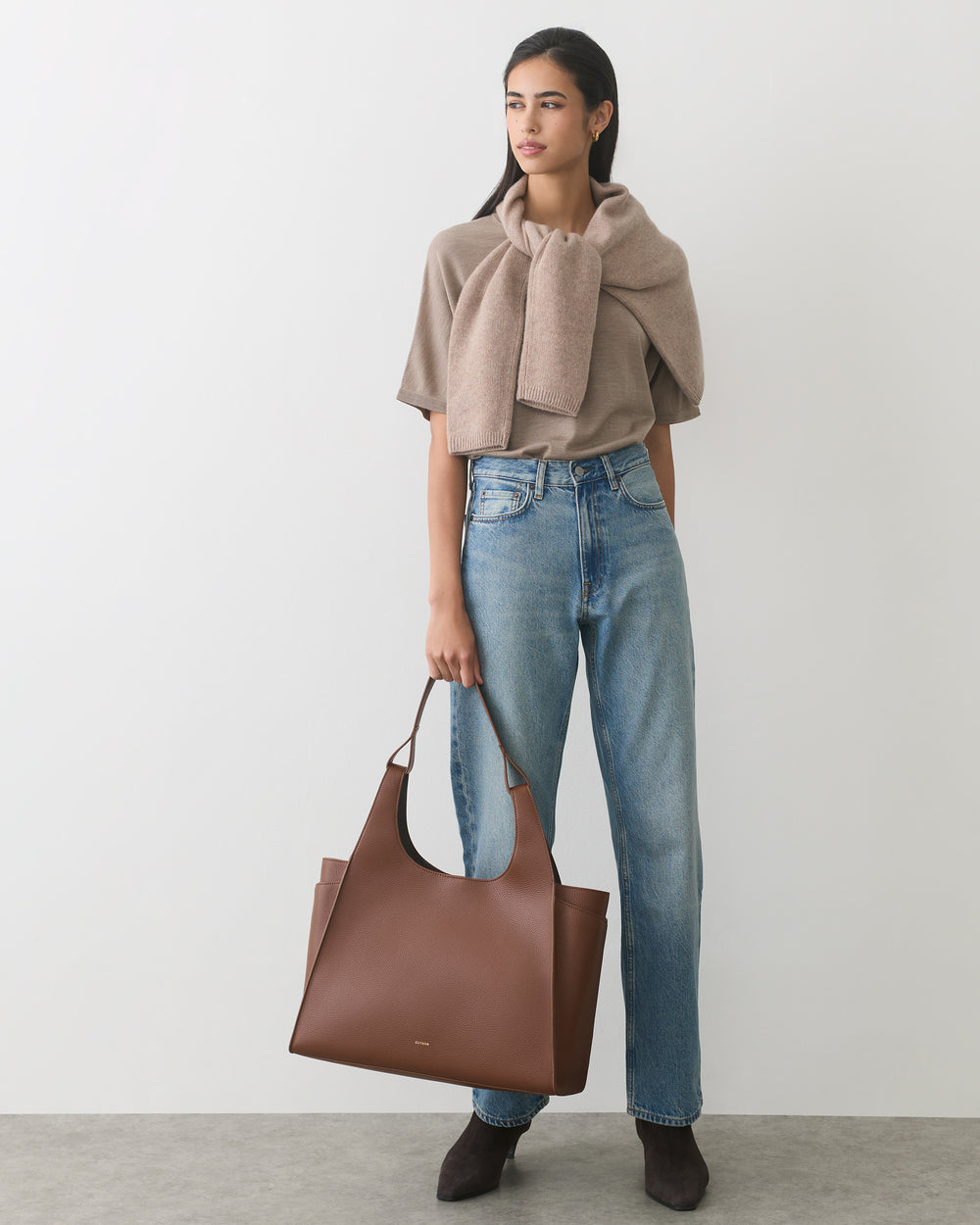 Person wearing jeans and top, carrying a large tote bag, stands against plain backdrop.