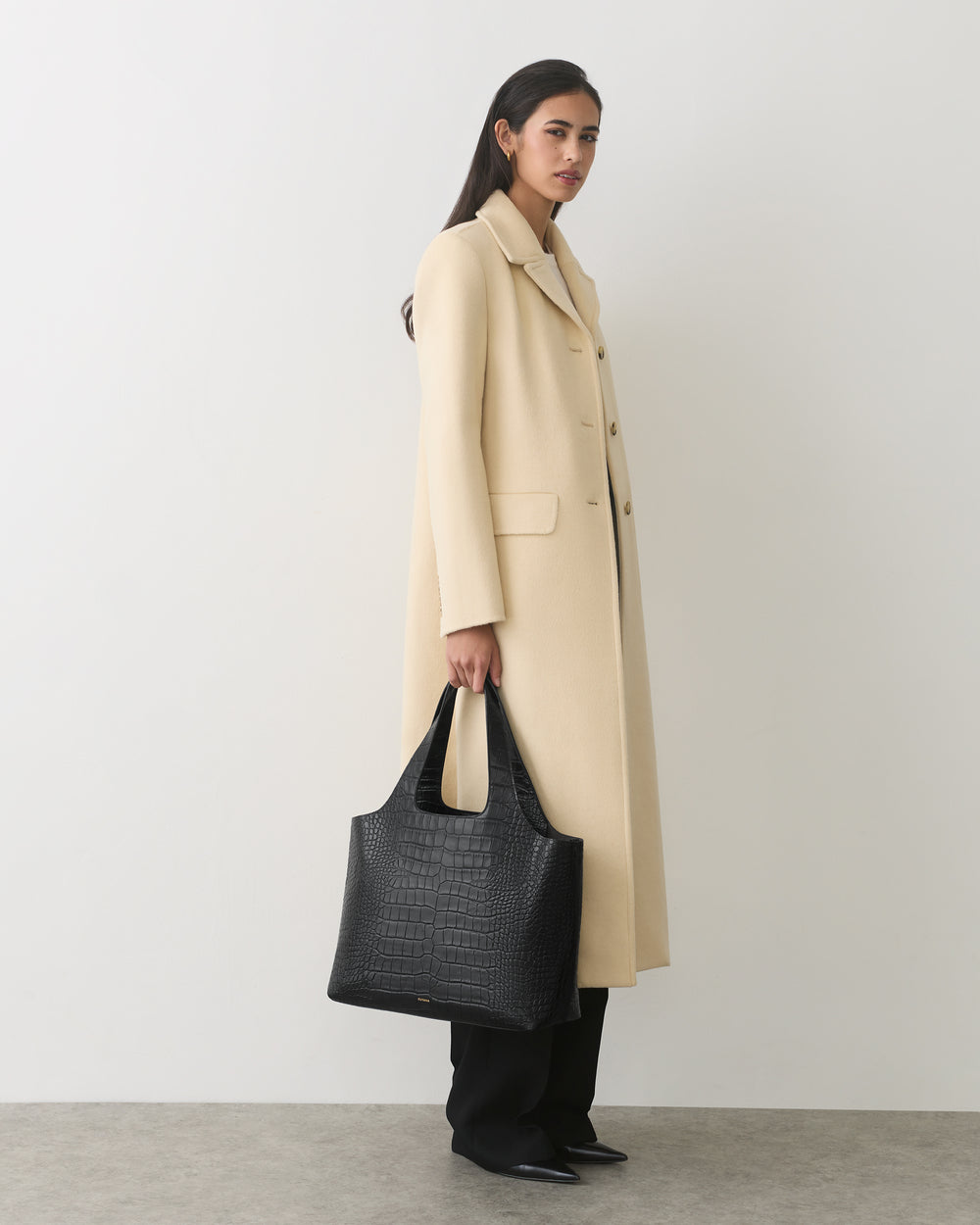 Person in long coat holding a croc-embossed leather bag standing against a plain backdrop