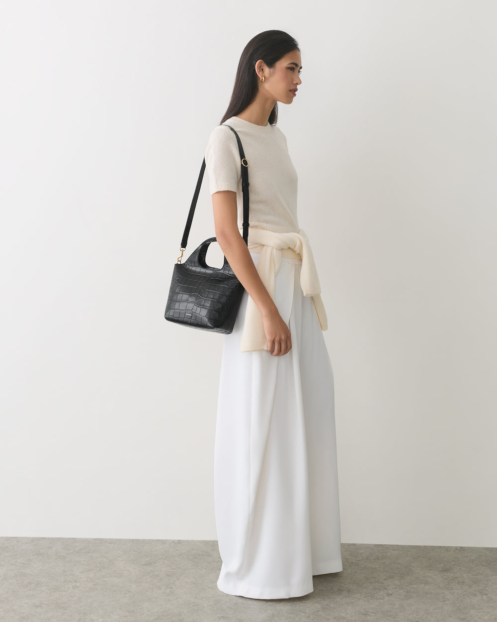 Person standing with a small croc-embossed leather bag over shoulder, wearing a tied sweater.