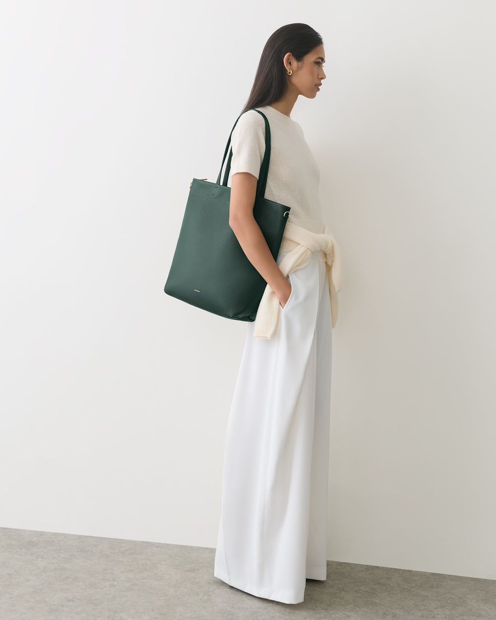 Person wearing a shirt and wide pants with a bag over shoulder standing against a plain backdrop.