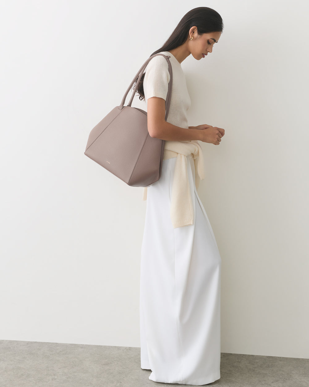Person standing sideways holding a handbag, wearing a long skirt and a top with tied sweater.