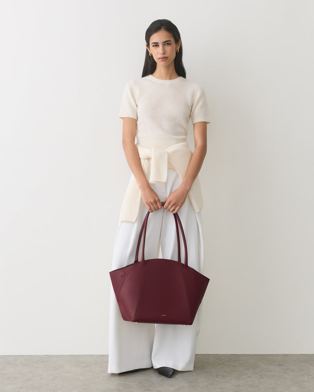Person holding large handbag wearing a short-sleeve top with tied sweater and wide-leg pants.