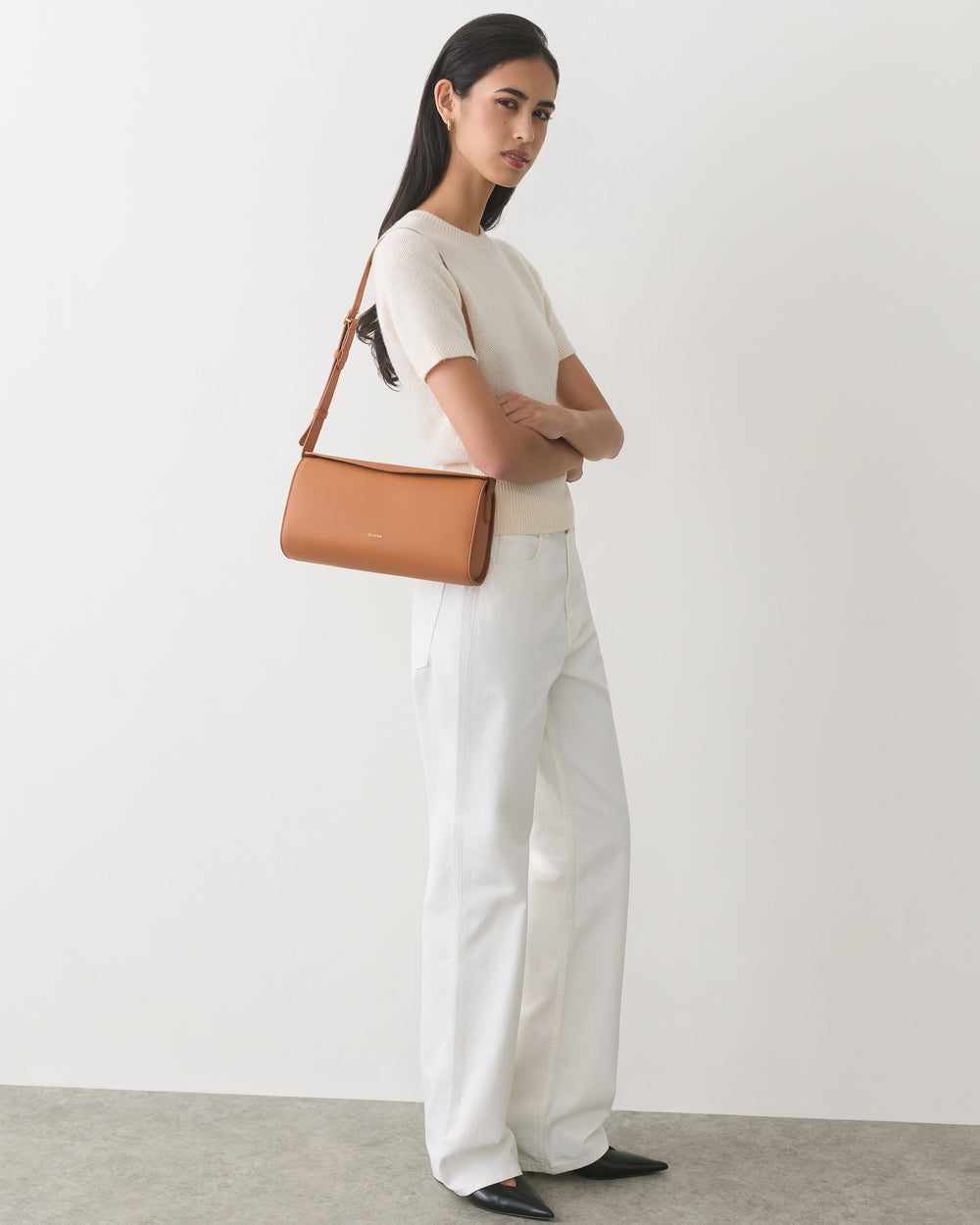 Person standing with arms crossed, wearing a short-sleeve top and pants, carrying a shoulder bag.