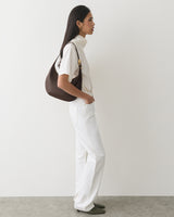Person in white clothes with shoulder bag standing against plain wall.