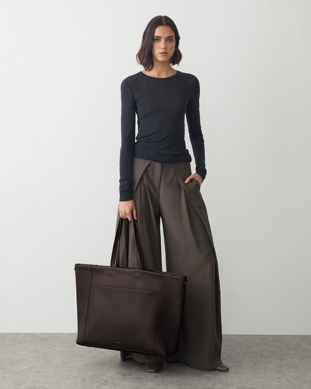 Person holding a large travel bag, wearing long-sleeve top and wide-leg pants.