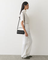 Person in white outfit holding a shoulder bag, standing on a plain background.
