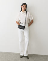 Person in white outfit, holding a black handbag, standing against a plain wall.