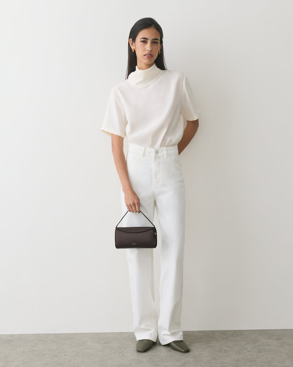 Person standing against plain wall holding a small handbag wearing a short-sleeve top and pants.