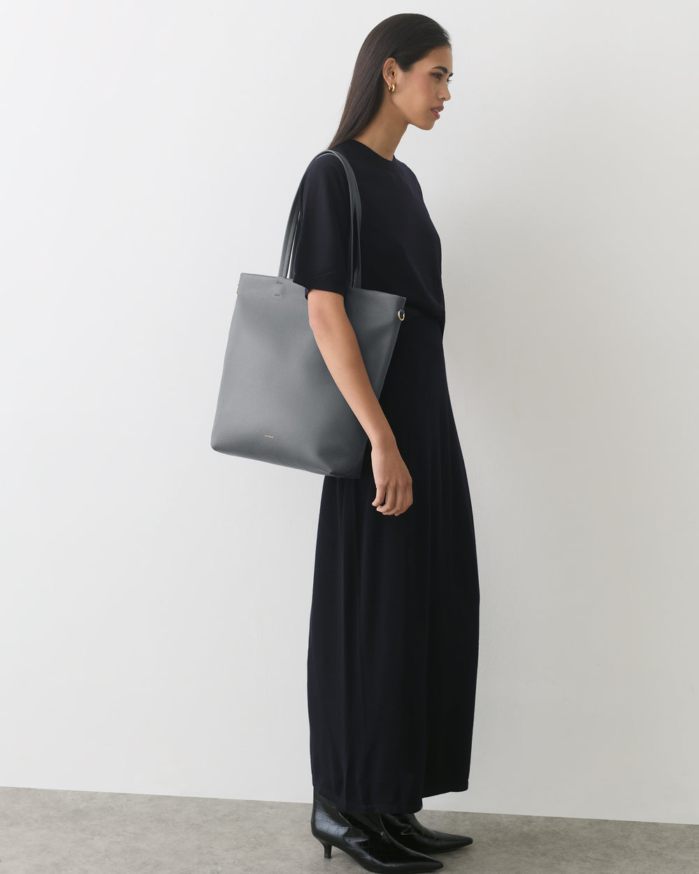 Person in dress holding tote bag, standing against plain wall.