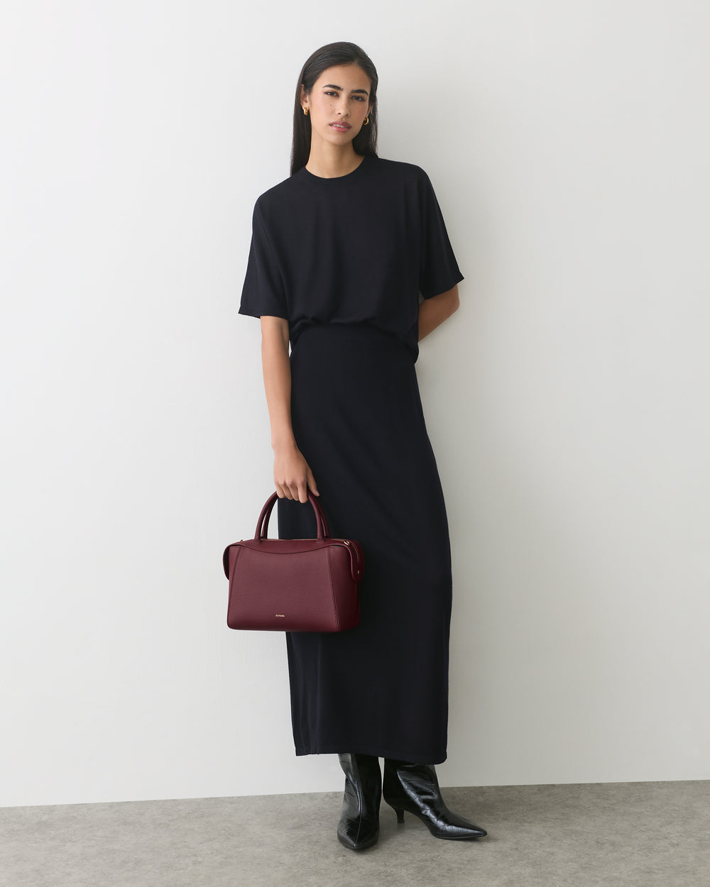 Person in long dress holding handbag, standing against a plain wall.