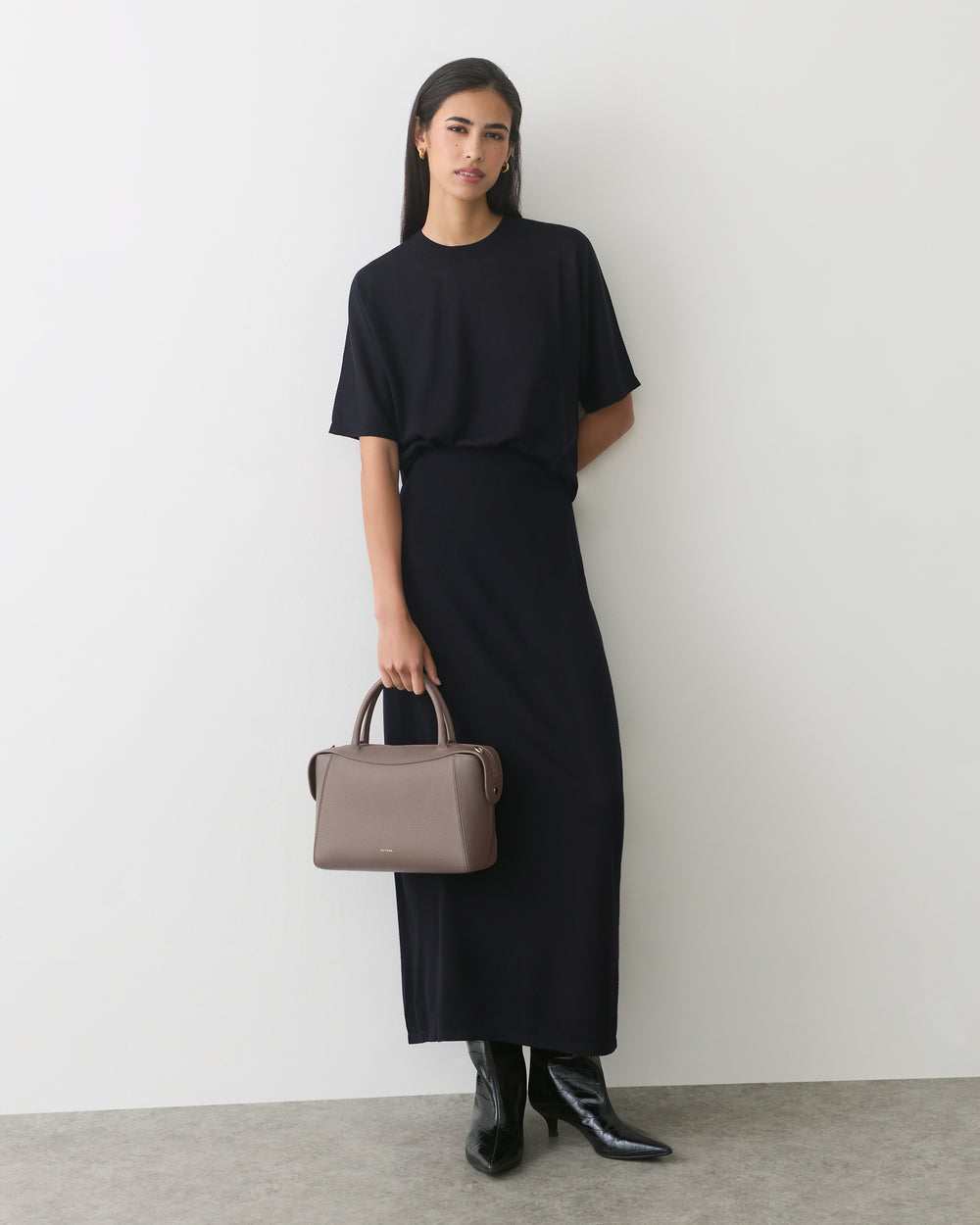 Model in dress holding a handbag, wearing boots, standing against white wall.