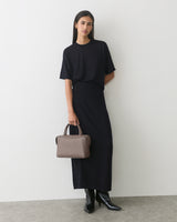 Model in dress holding a handbag, wearing boots, standing against white wall.