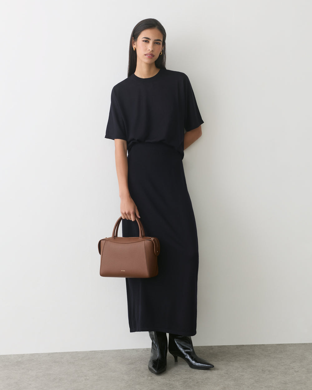 Person in long dress holds handbag, wearing boots, stands against plain backdrop.