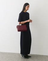 Person in long dress holding handbag, standing against plain wall.