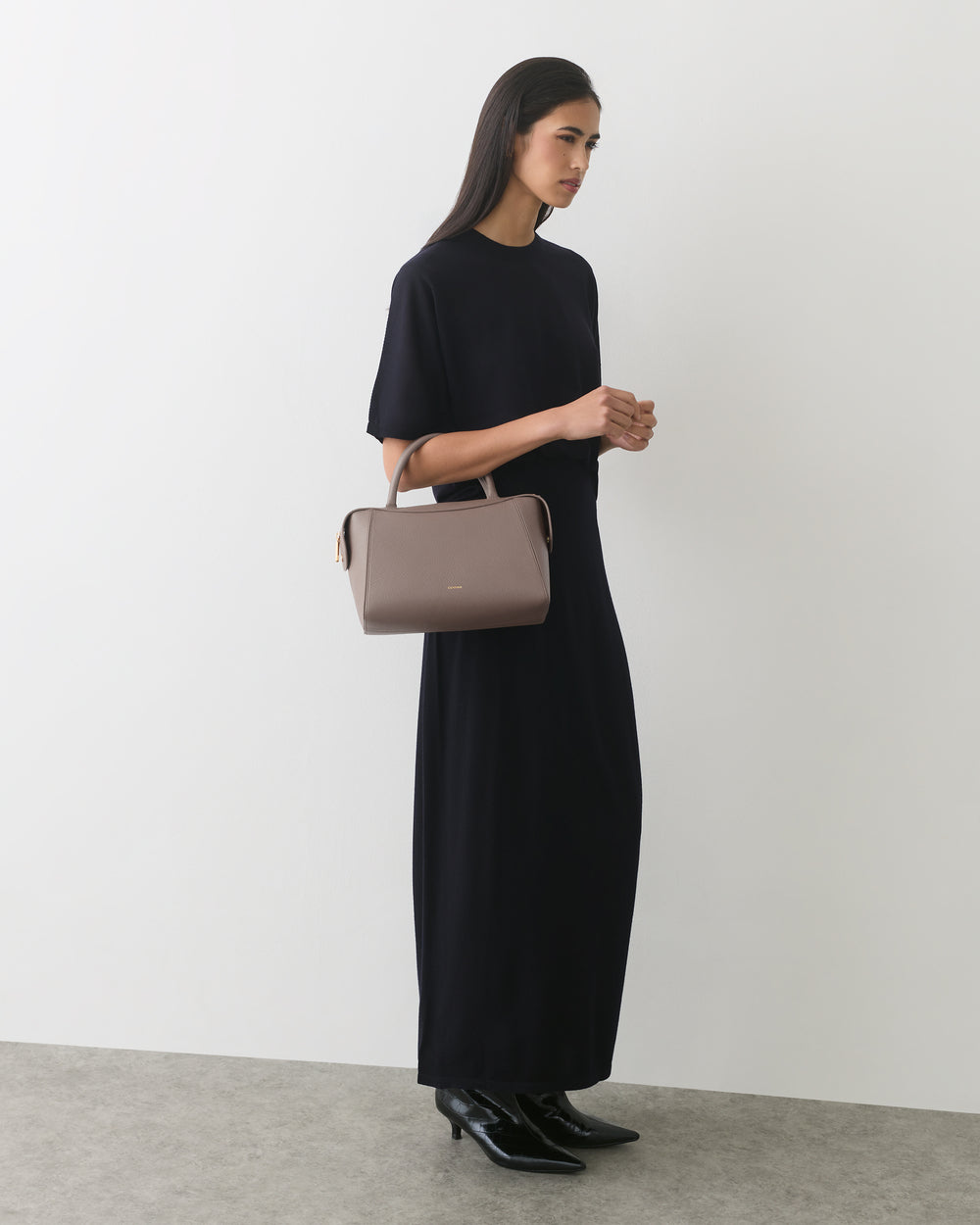 Person wearing long dress holding handbag, standing against plain wall.