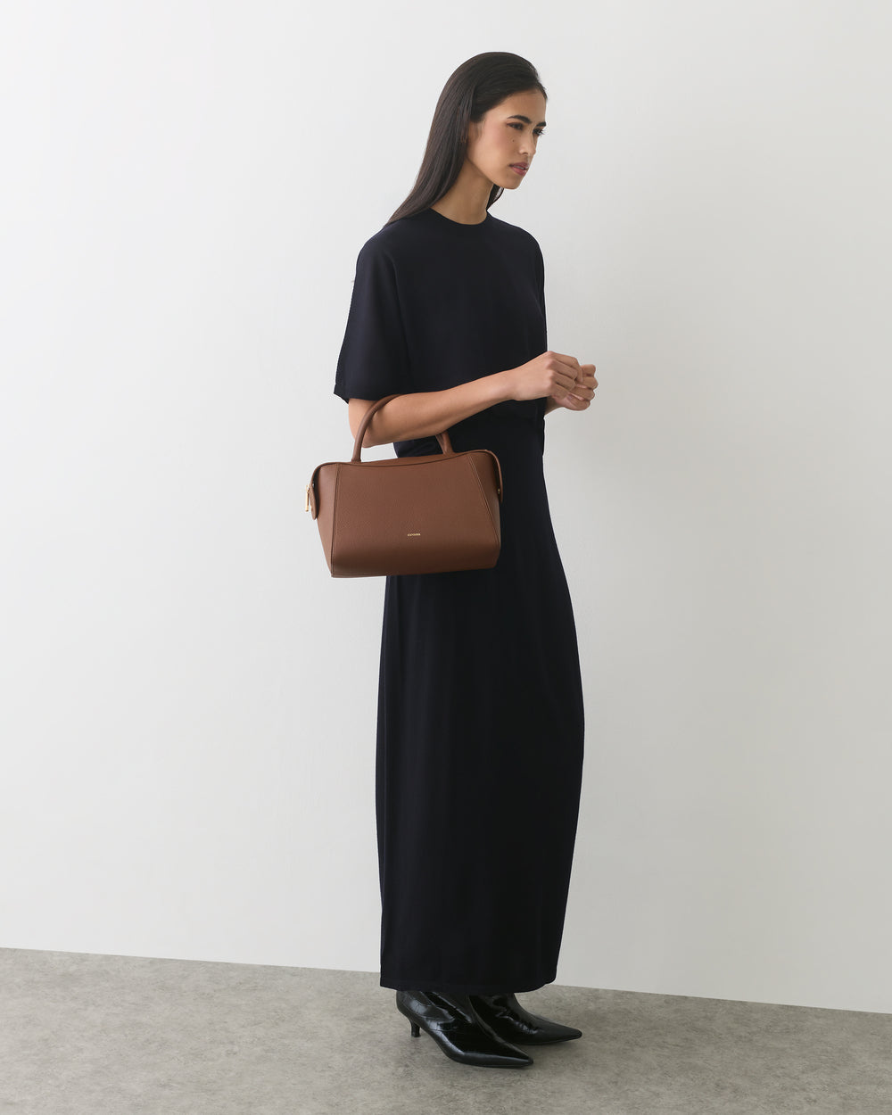 Person in long dress holding a handbag, standing against a plain backdrop.
