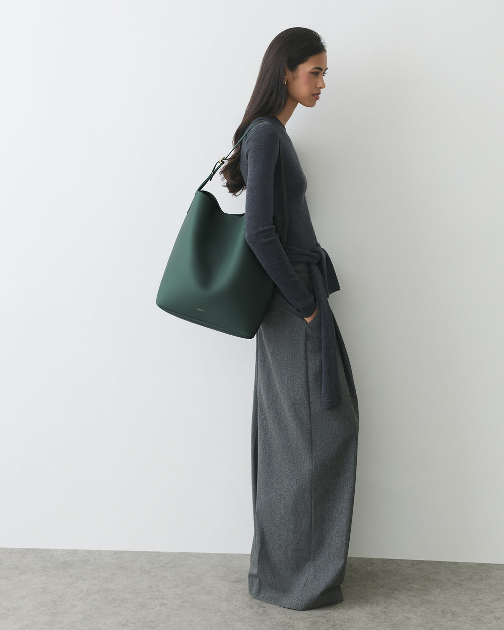 Person in a long outfit carrying a large shoulder bag standing against a plain wall.