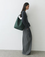 Person in a long outfit carrying a large shoulder bag standing against a plain wall.