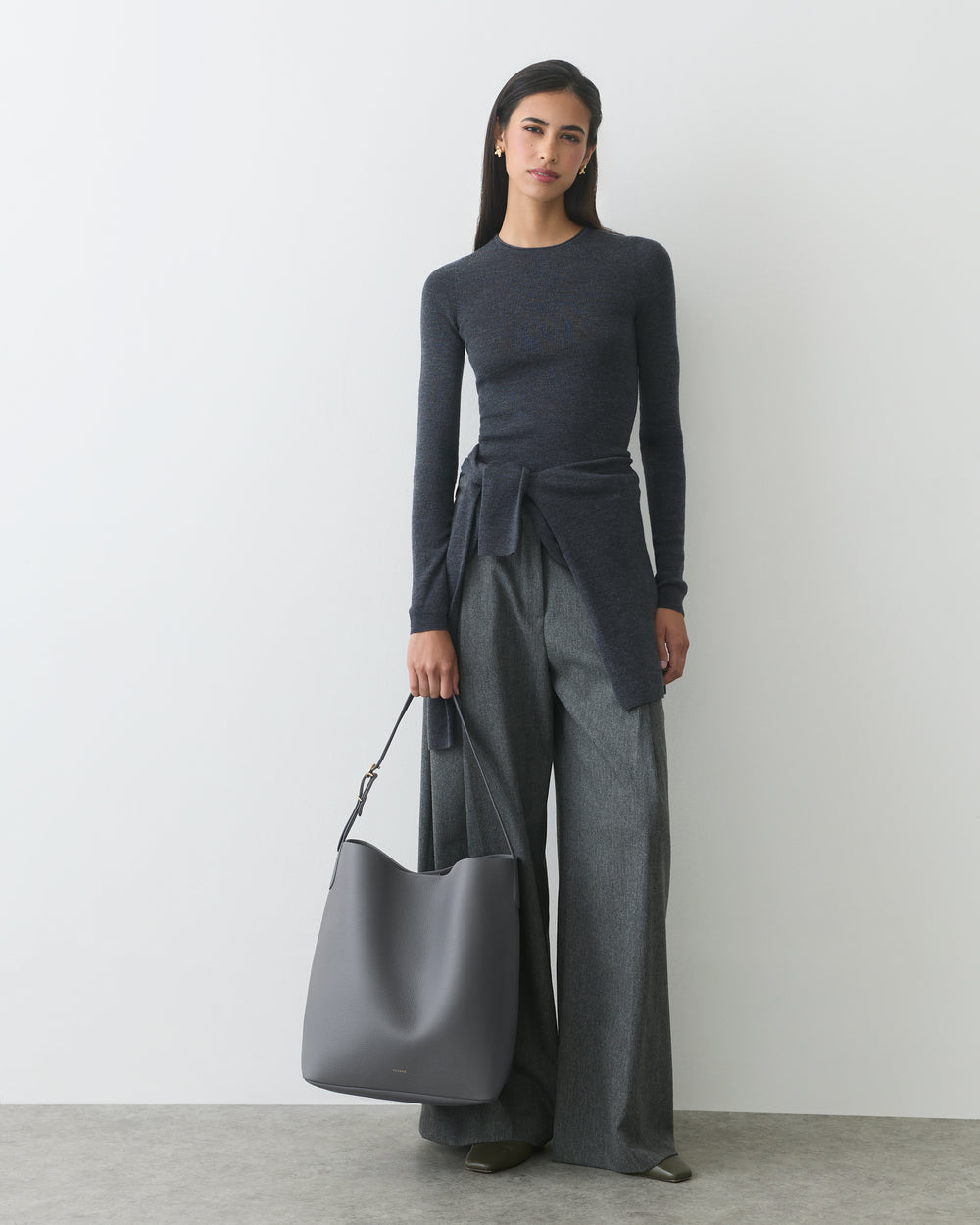A person stands holding a large shoulder bag, wearing a long-sleeve top and wide-leg trousers.