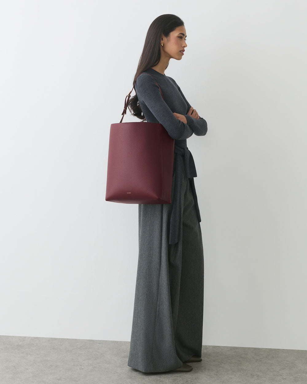 Person standing with arms crossed, holding large tote bag, wearing long pants and long-sleeve top