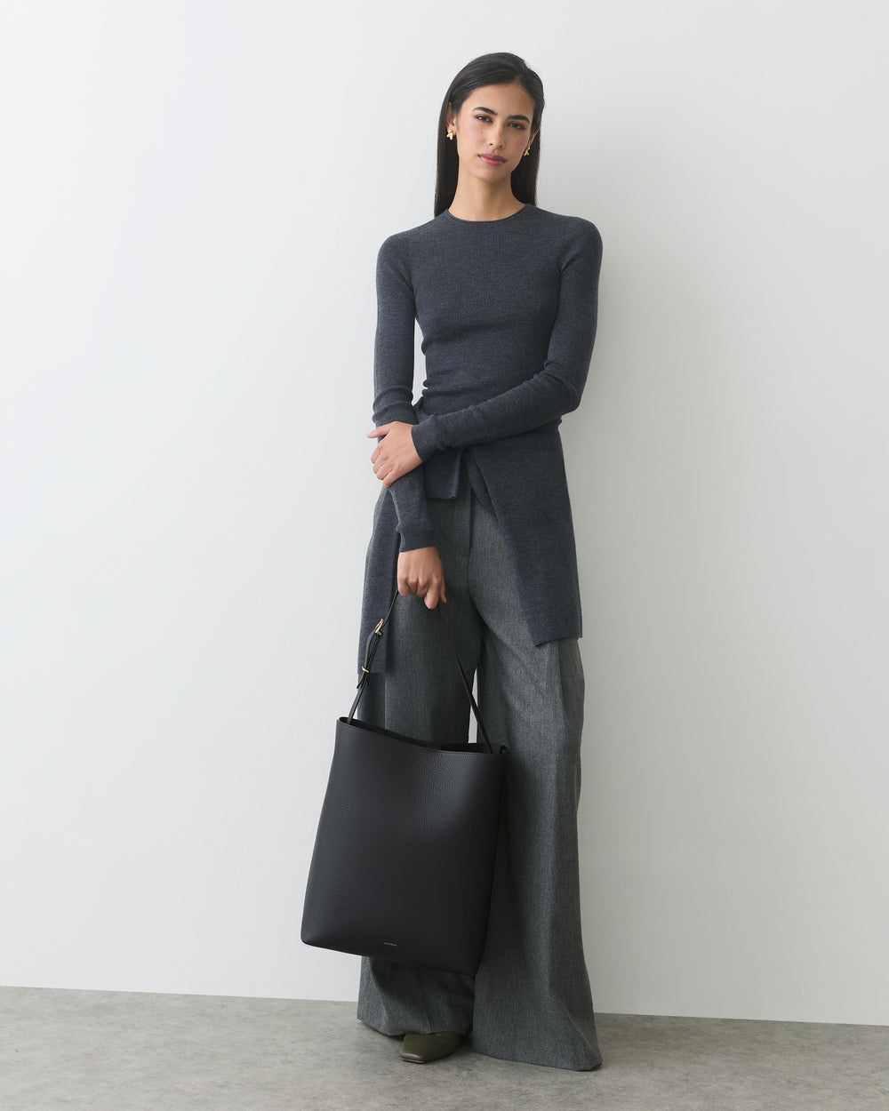 Woman in long sleeve top and wide leg pants holding a tote bag standing against a white wall