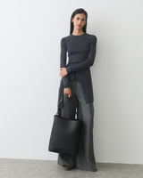 Woman in long sleeve top and wide leg pants holding a tote bag standing against a white wall