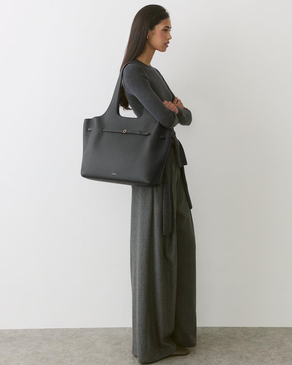 Person in long dress holding large shoulder bag standing against plain background.