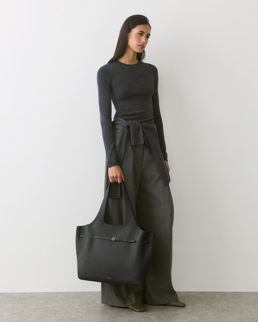 Person with long sleeves, wide pants, holding a large handbag, standing against plain backdrop.