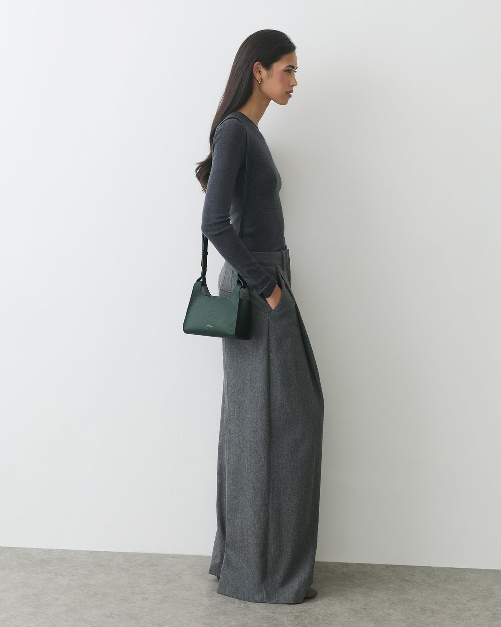Person in long-sleeve top and wide-leg pants, holding handbag, standing against a wall.