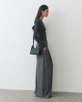Person in long-sleeve top and wide-leg pants, holding handbag, standing against a wall.