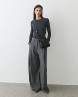 Person wearing long-sleeve top, wide-leg trousers, crossbody bag, standing against plain backdrop.