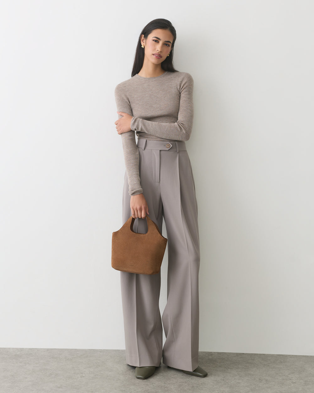 Woman in long-sleeve top and wide-leg pants holding a handbag, standing against a plain wall.