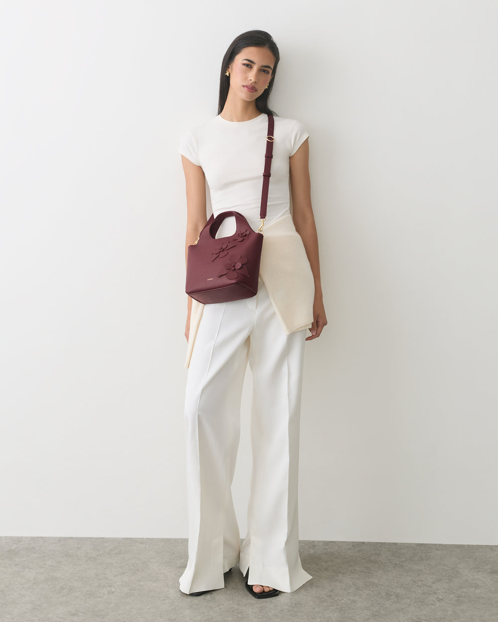 Person standing with a shoulder bag with a floral detail, wearing a top and wide-leg pants.
