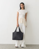 Person standing holding a large handbag wearing a short-sleeve top and pants.