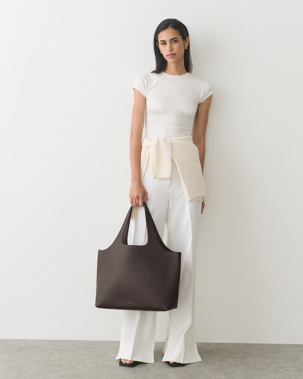Person stands against a plain wall holding a large handbag and wearing a casual outfit.