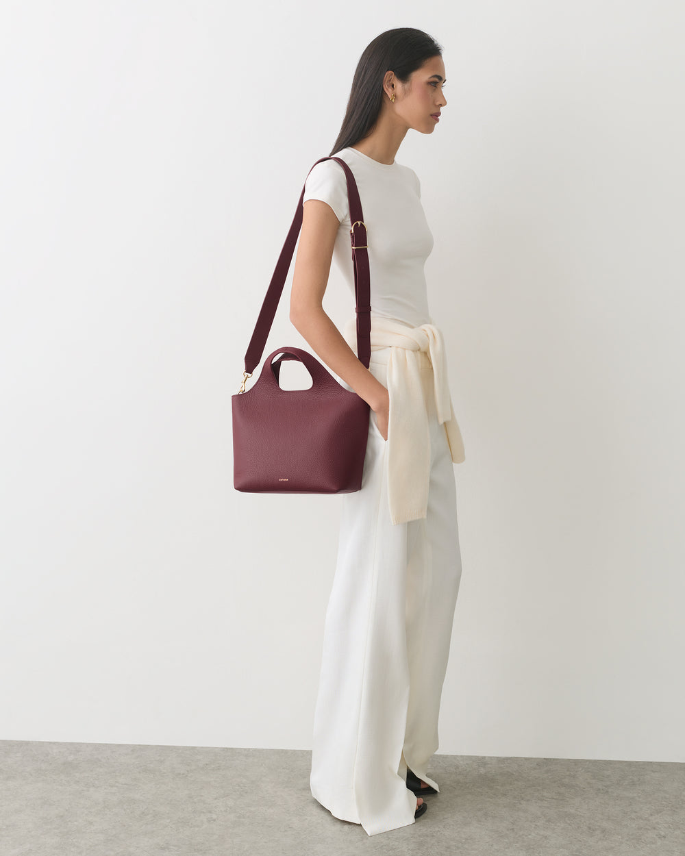 Person standing sideways with a bag on shoulder, wearing a tied sweater and wide-leg pants.