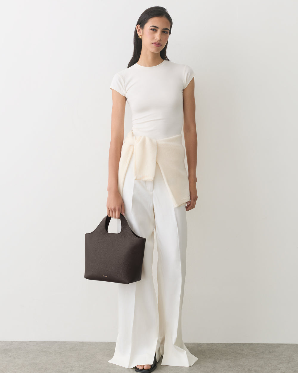 Person wearing a shirt and wide-leg pants, holding a handbag, with a sweater tied around waist.