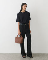Person wearing a t-shirt and pants holding a handbag standing against a plain background.