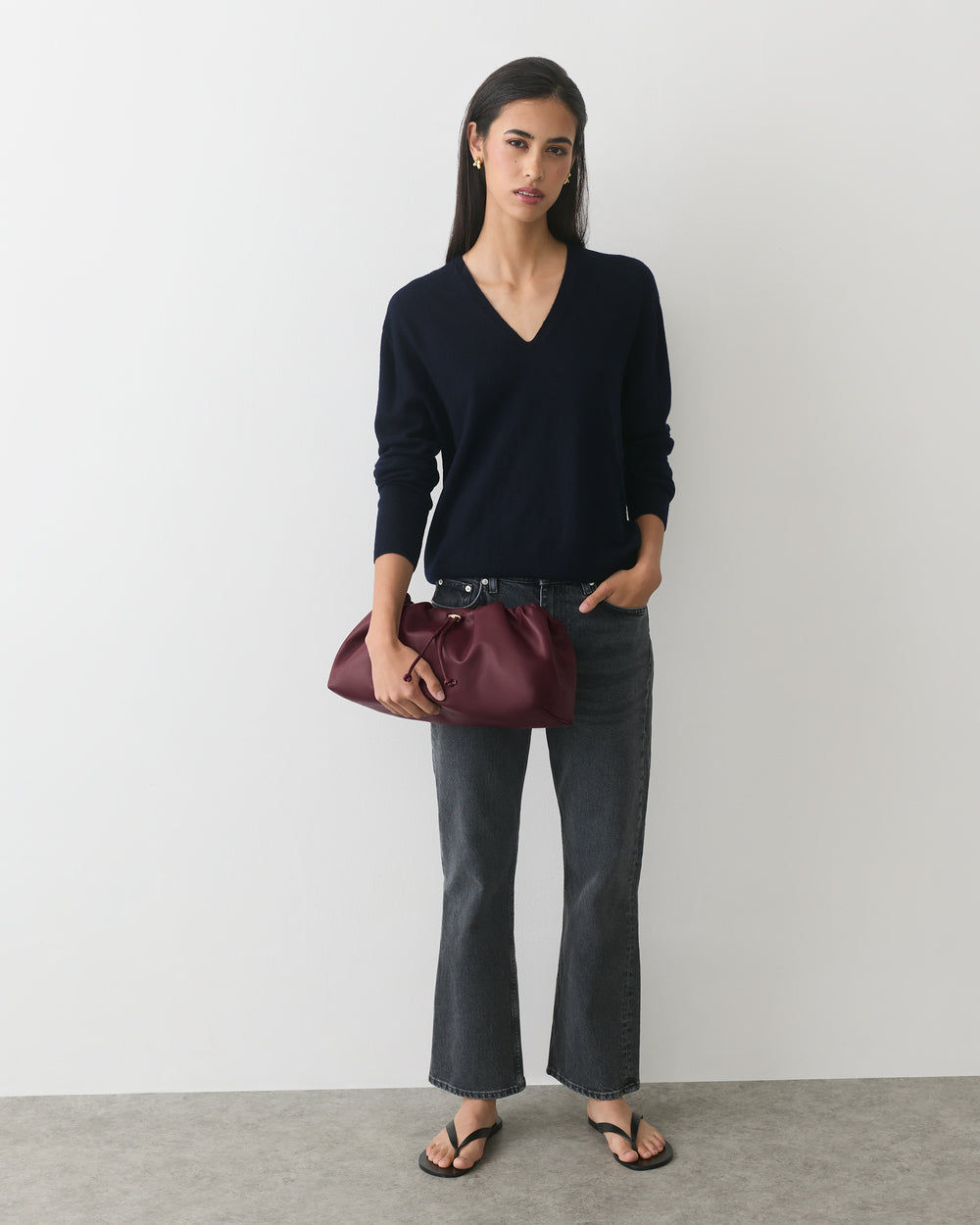Person holding a handbag, wearing a sweater, jeans, and sandals standing in front of a plain wall.