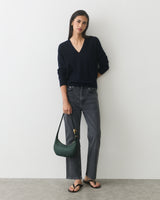 Person wearing a sweater and jeans, holding a handbag, standing against a plain background.