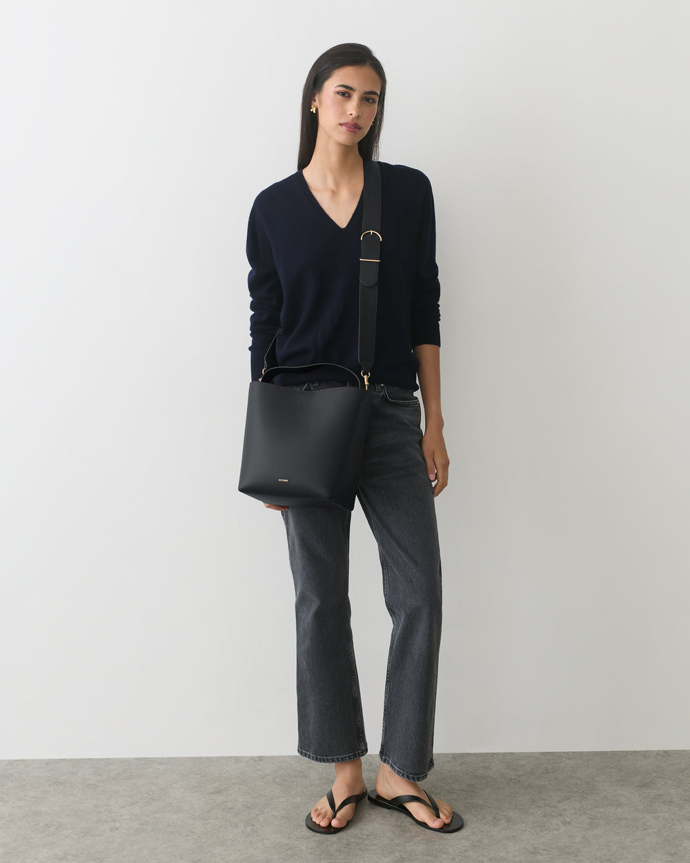 Person wearing a sweater and jeans, holding a shoulder bag, standing in a minimalist room.