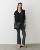 Person wearing a sweater and jeans, holding a shoulder bag, standing in a minimalist room.